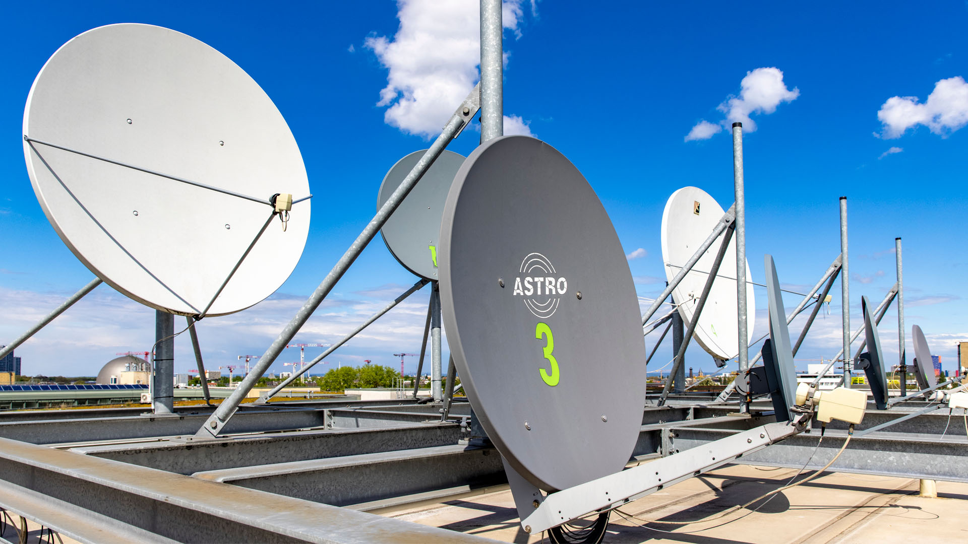 Reihe professioneller Satelliten-Uplink-Schüsseln auf einem Dach vor blauem Himmel mit Baukränen im Hintergrund