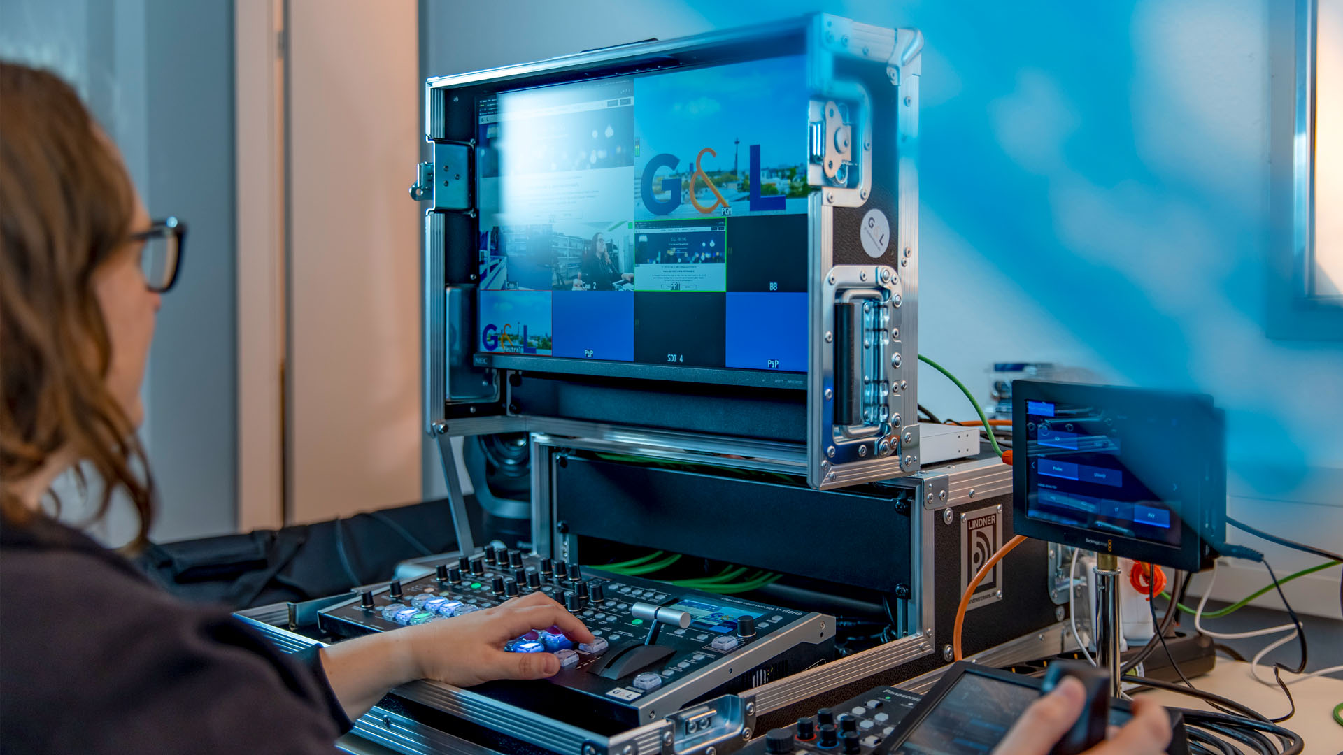 Techniker bedient ein mobiles Broadcast-Produktionsrack mit Multiview-Monitor, der G&L-Branding und Kamerabilder zeigt, Hände an einem Audio-/Video-Mischpult